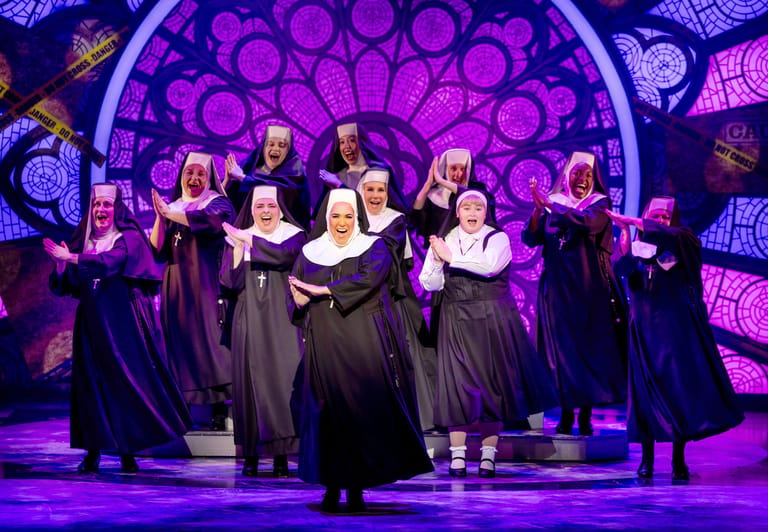 Choir members in dark robes sing together on stage beneath glowing purple circular backdrop lighting.