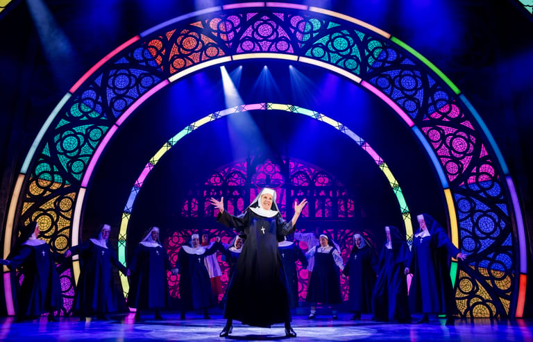 Performer in dark robes stands centre stage beneath an ornate illuminated archway with ensemble cast in purple stage lighting.
