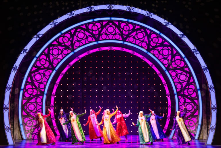 Performers in flowing gowns stand beneath a glowing arch of purple and magenta lights on a theatrical stage.