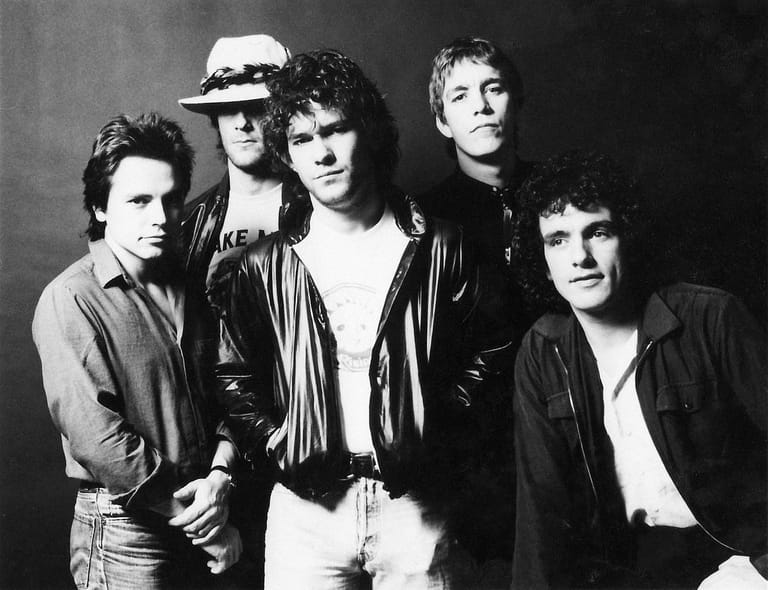 Five musicians in 1970s-style clothing pose against a dark background in black and white.