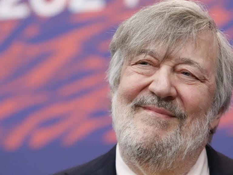 Older man with grey beard and hair smiling at a public event with coloured backdrop.