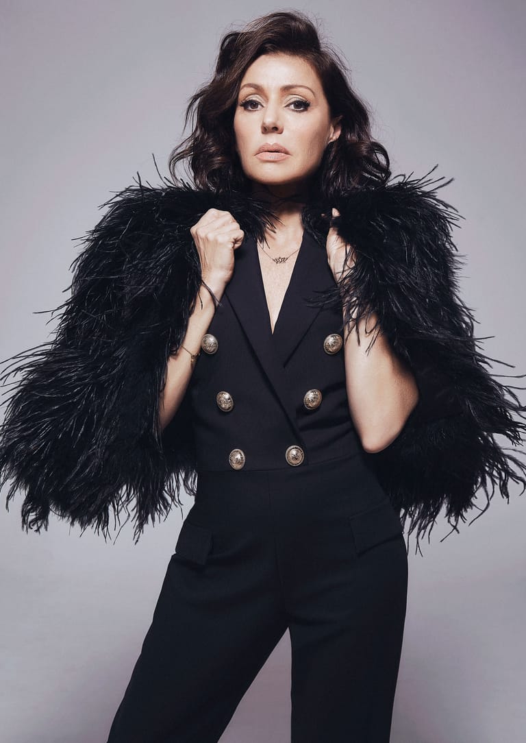 Woman in black double-breasted blazer with black feathered shoulders against neutral grey background.