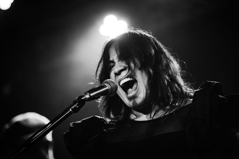 Performer singing passionately at a microphone beneath dramatic stage lighting in black and white.