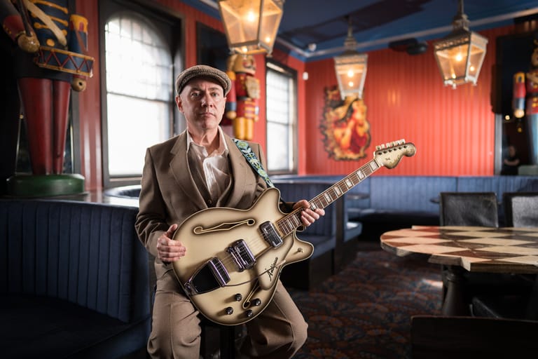 Musician holding an electric guitar in a vintage-styled red and blue interior room with warm lighting.