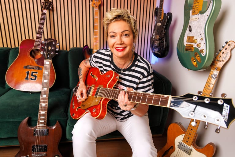 Musician in striped shirt holding red semi-acoustic guitar surrounded by instruments on wooden wall panelling.