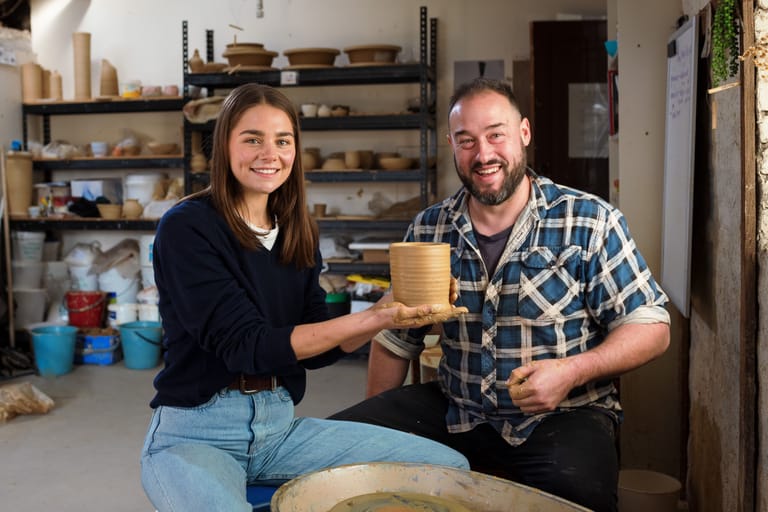 Two ceramicists display handmade pottery in a studio workshop lined with shelves of work.