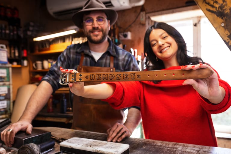 Man in cowboy hat and woman in red dress hold a wooden guitar together in a workshop setting.