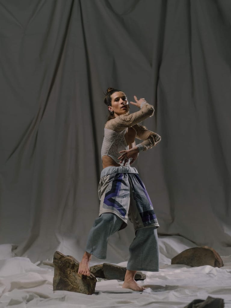 Shirtless dancer posed dynamically against grey draped fabric with rocks scattered on white floor.