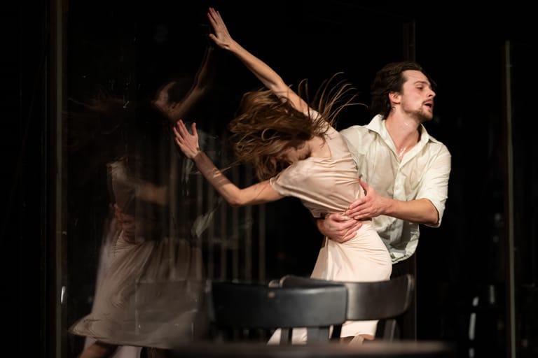 Two dancers in period costume perform an intimate theatrical scene under stage lighting, one seated and one moving expressively beside him.