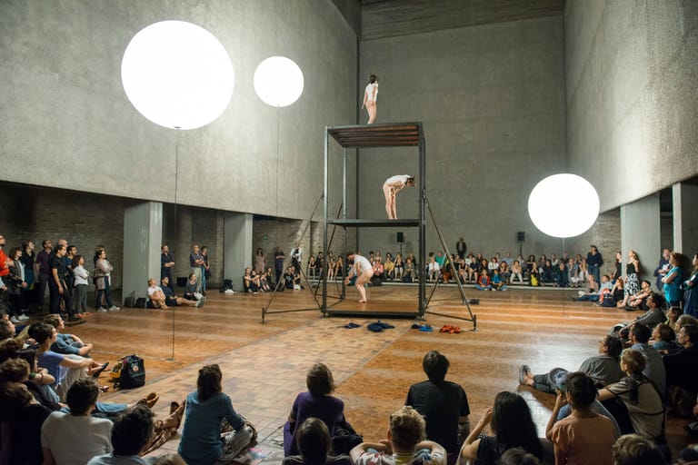Performer on a raised platform suspended above a wooden floor, surrounded by seated audience members in a minimalist concrete theatre space.