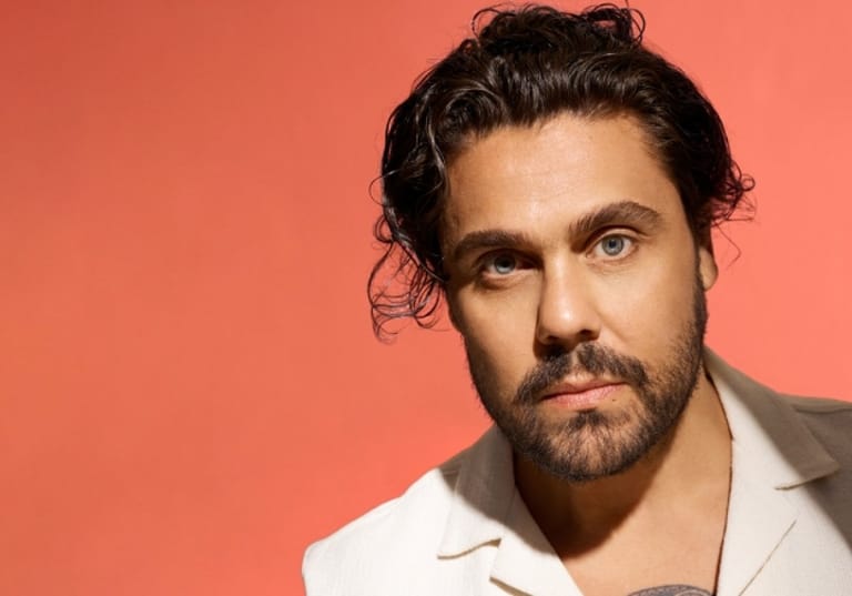 Man with dark hair and beard wearing a white shirt against a coral-red background.