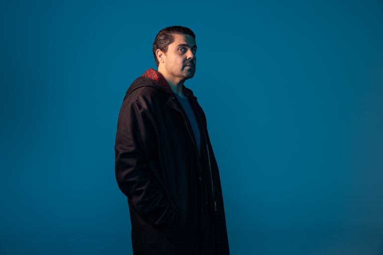 Man in dark jacket standing in profile against blue backdrop.