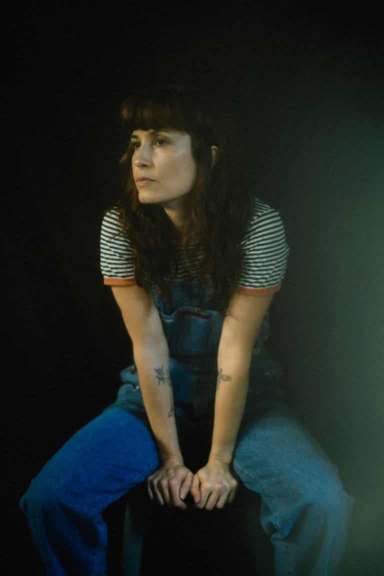 Woman with dark hair and fringe wearing striped shirt and blue overalls, seated against dark green background.