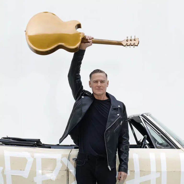 Man in black leather jacket holding a gold acoustic guitar aloft against a white sky.