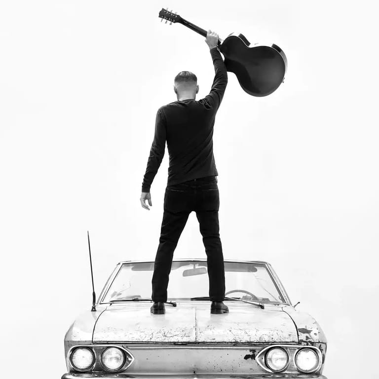 Man in black suit stands on vintage car roof holding acoustic guitar aloft against white background.