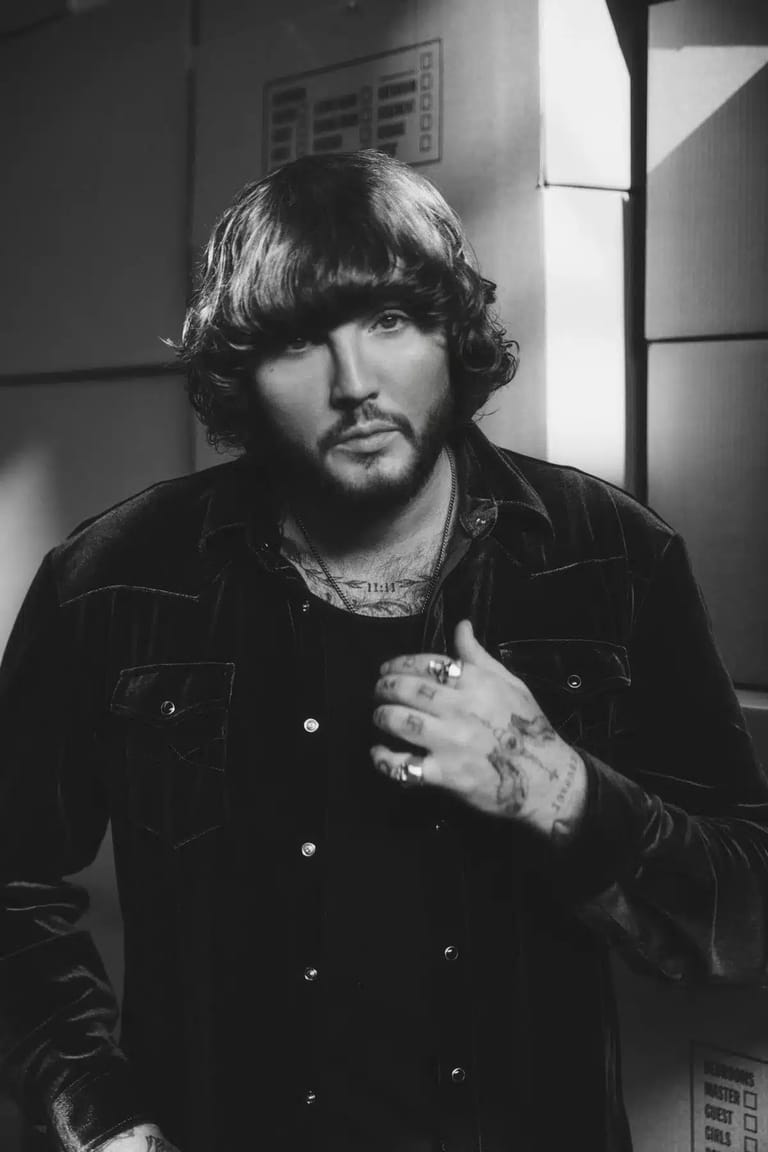 Man with shoulder-length dark hair wearing a black jacket in a black and white studio portrait.