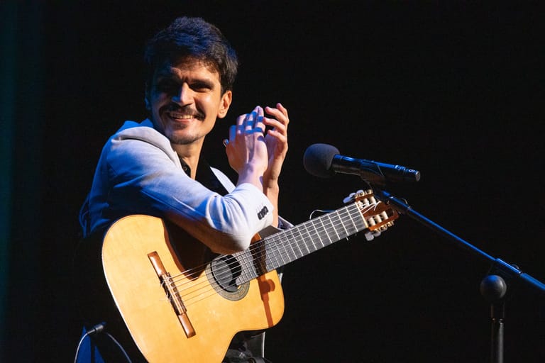Musician performing on stage with acoustic guitar and microphone, smiling at audience.