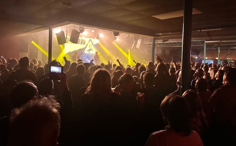 Crowd of concert-goers with raised hands and phone cameras facing a brightly lit stage with yellow lighting.