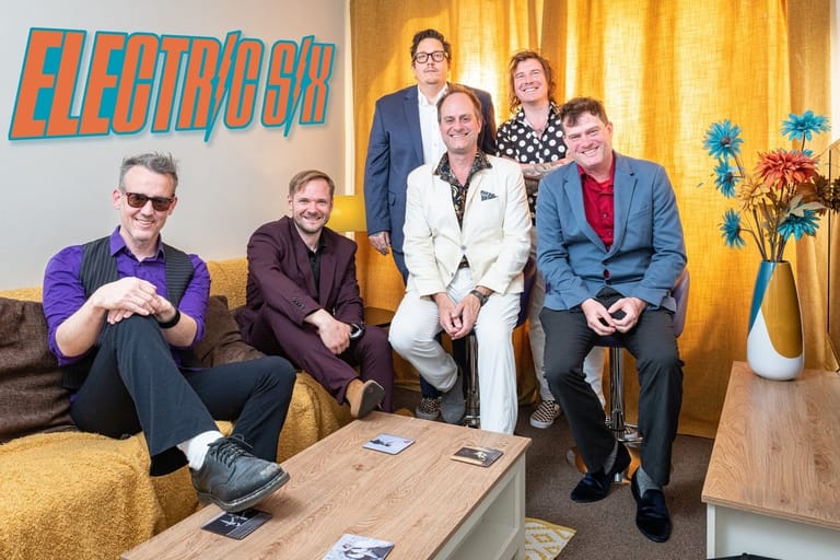 Five band members posed in a living room setting with colourful "Electric 6" signage and warm yellow curtains behind them.