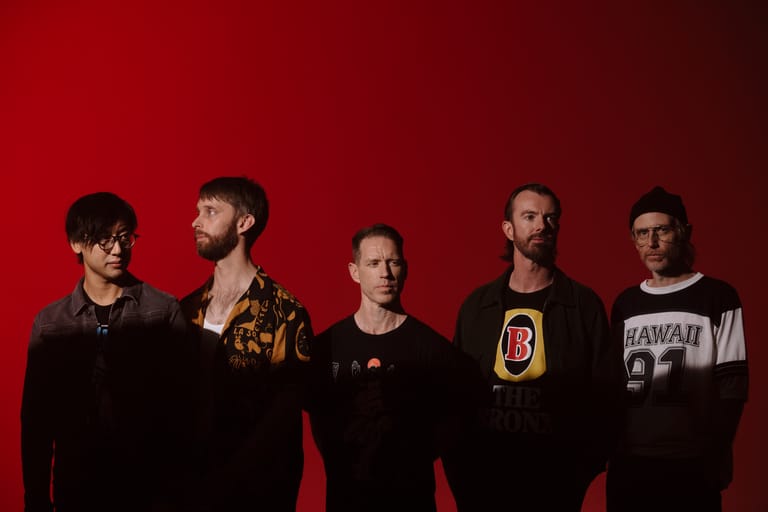 Five band members stand against a red backdrop in dark clothing and casual poses.