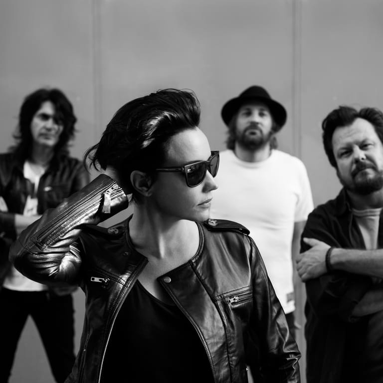 Four musicians in casual attire pose against a white backdrop in black and white, with the front figure wearing sunglasses and a leather jacket.
