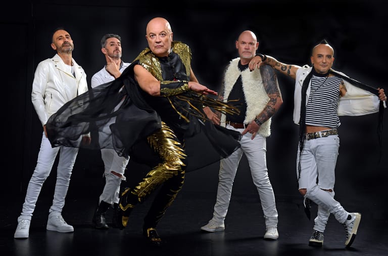 Five performers in white and gold clothing pose dynamically on a dark stage during a theatrical production.