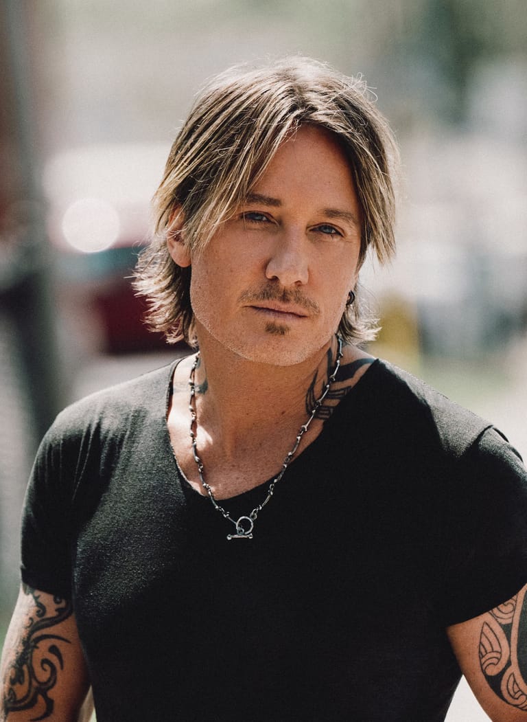 Man with shoulder-length blonde hair and arm tattoos wearing a black shirt and chain necklace outdoors.