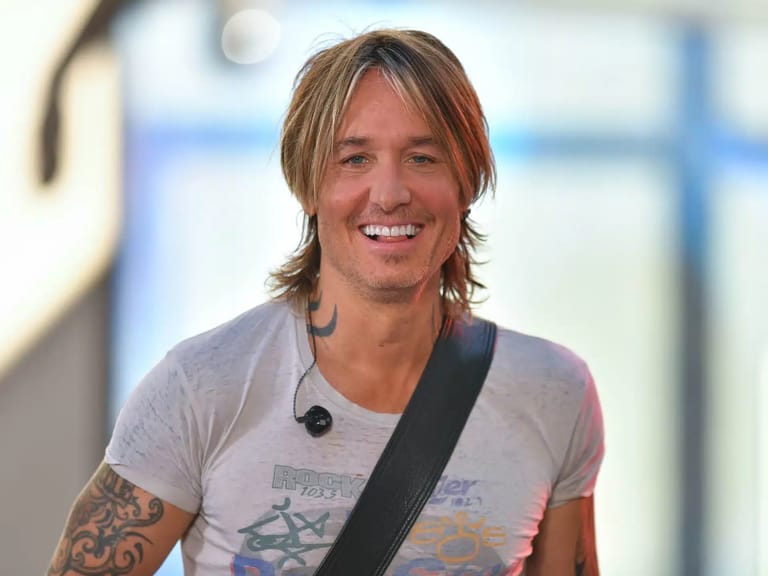 Man with shoulder-length blonde hair and arm tattoo wearing microphone headset at indoor venue.