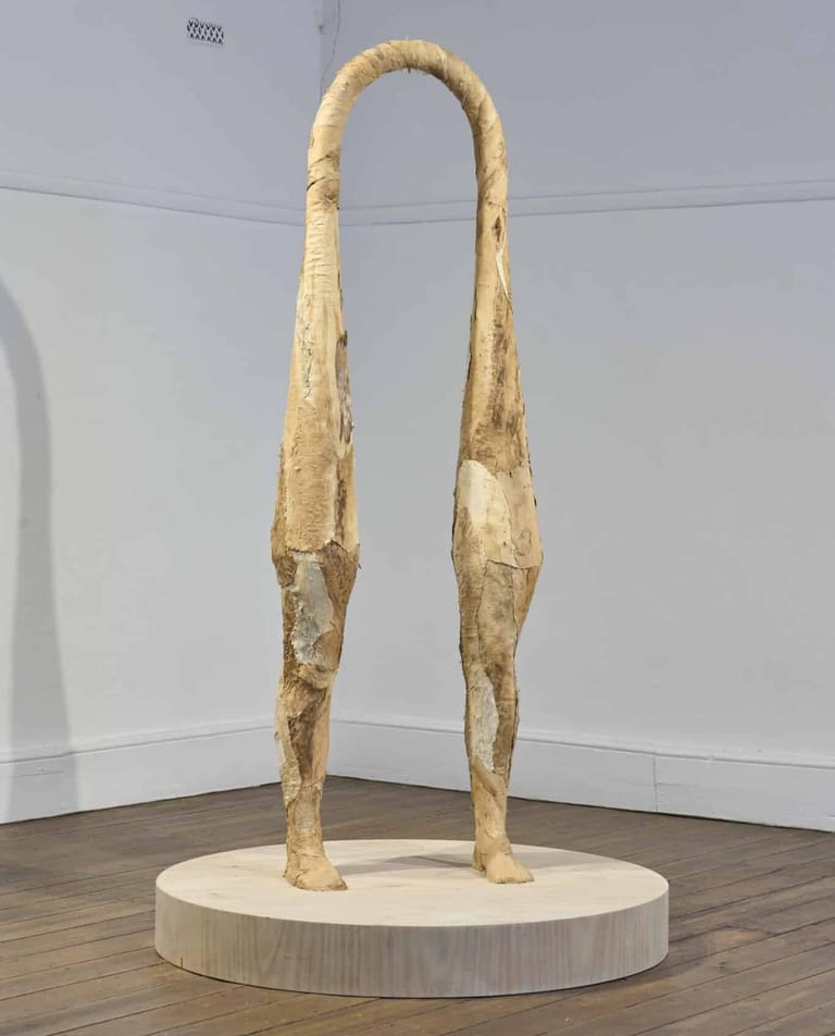 Tall wooden sculpture with two elongated legs joined at a curved arch, displayed on a circular platform in a gallery space.