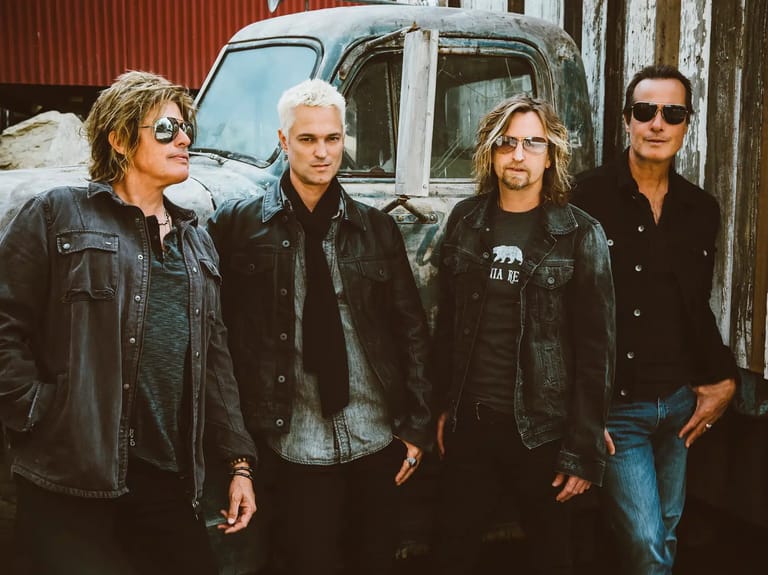 Four musicians in dark leather jackets stand against a weathered corrugated iron wall and vintage vehicle.