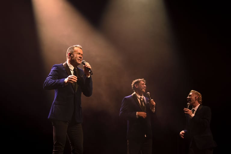 Three men in formal suits perform with microphones under dramatic stage lighting.