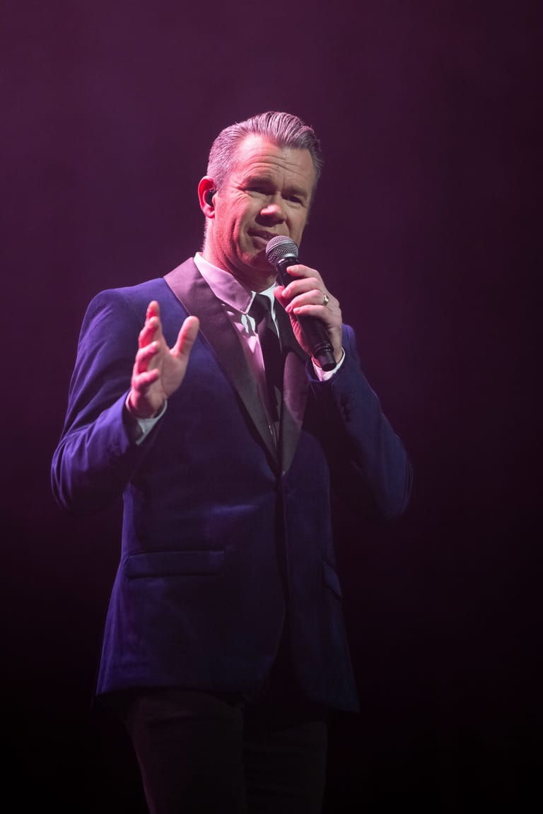 Man in dark suit speaking into microphone under purple stage lighting, gesturing expressively.