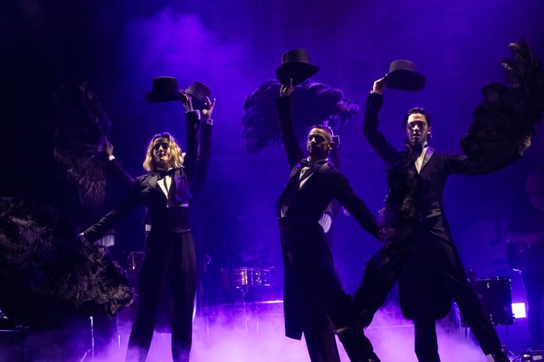 Multiple performers in dark suits stand on a stage bathed in purple and blue lighting during a live music performance.