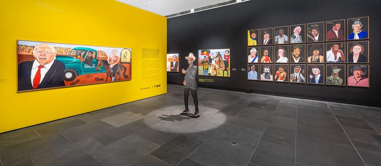 Gallery visitor standing in exhibition space between yellow wall with portrait imagery and black wall displaying multiple framed photographs.