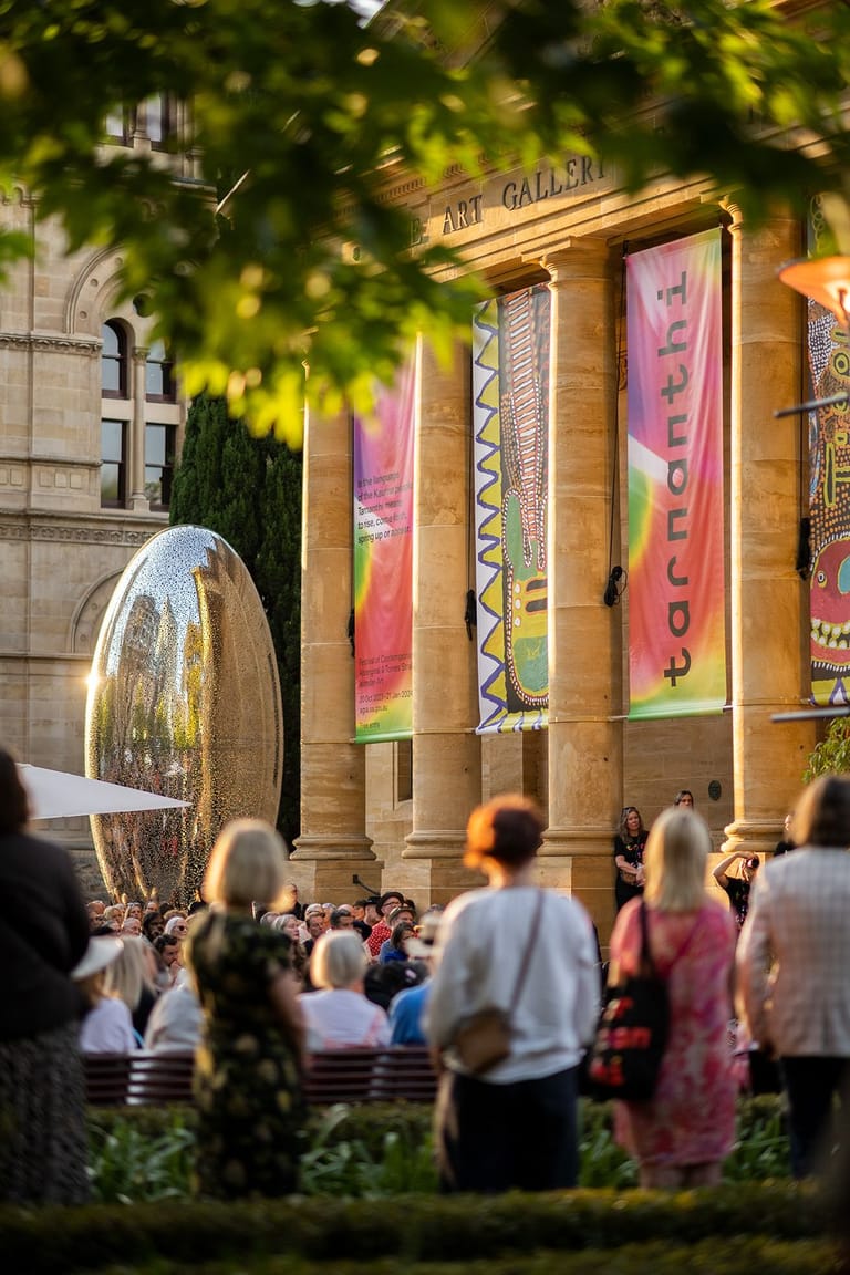 Crowds gather outside an art gallery displaying large colourful banners beneath leafy green foliage.