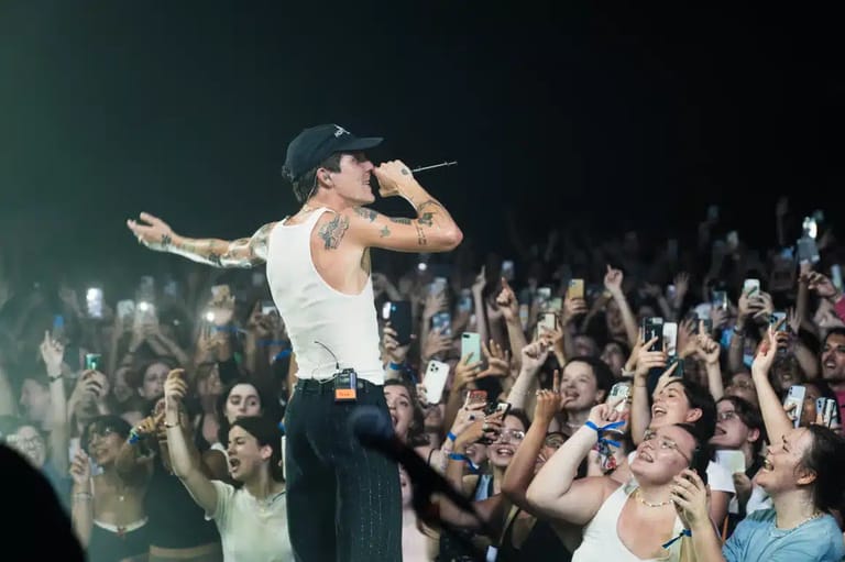 Performer in white singlet singing to a crowd of enthusiastic audience members holding phones aloft.