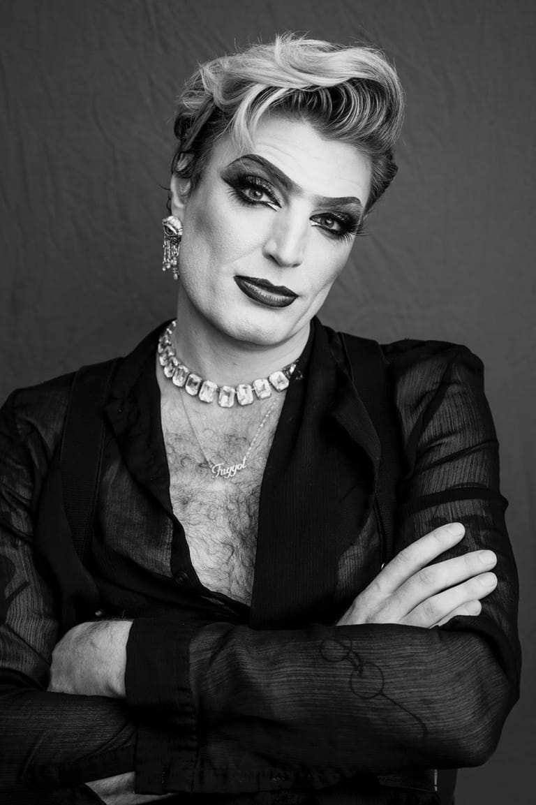 Person in black blazer and statement jewellery poses with arms crossed in dramatic black and white portrait.