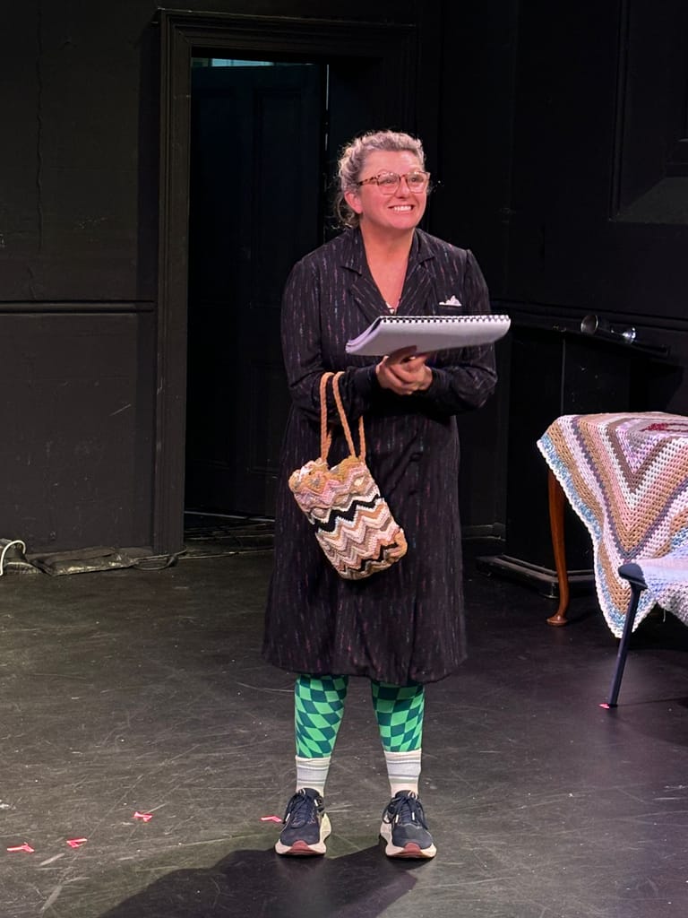 Performer in pinstriped coat and patterned socks holds a large prop knife on stage during a theatrical production.