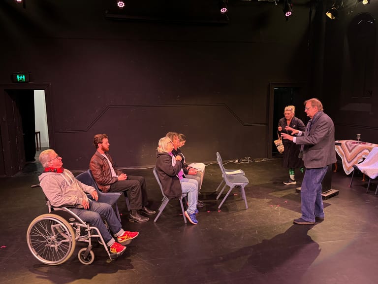 Five people seated in a circle on a darkly lit theatre stage, including one in a wheelchair, engaged in conversation.
