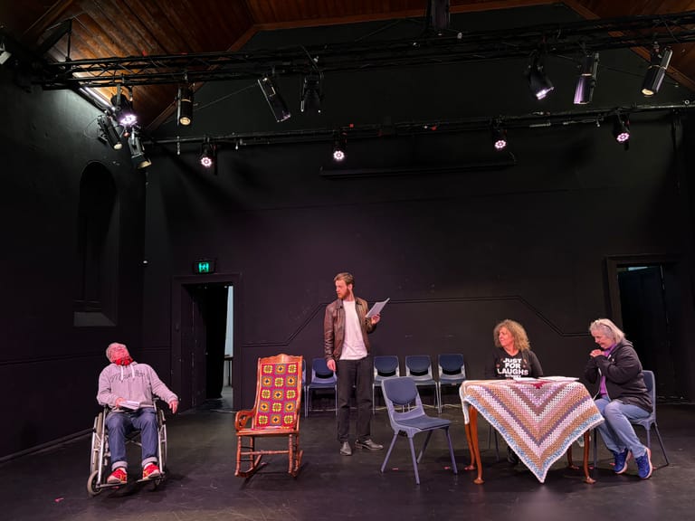 Four people engaged in theatrical performance on a dimly lit stage with chairs and props.