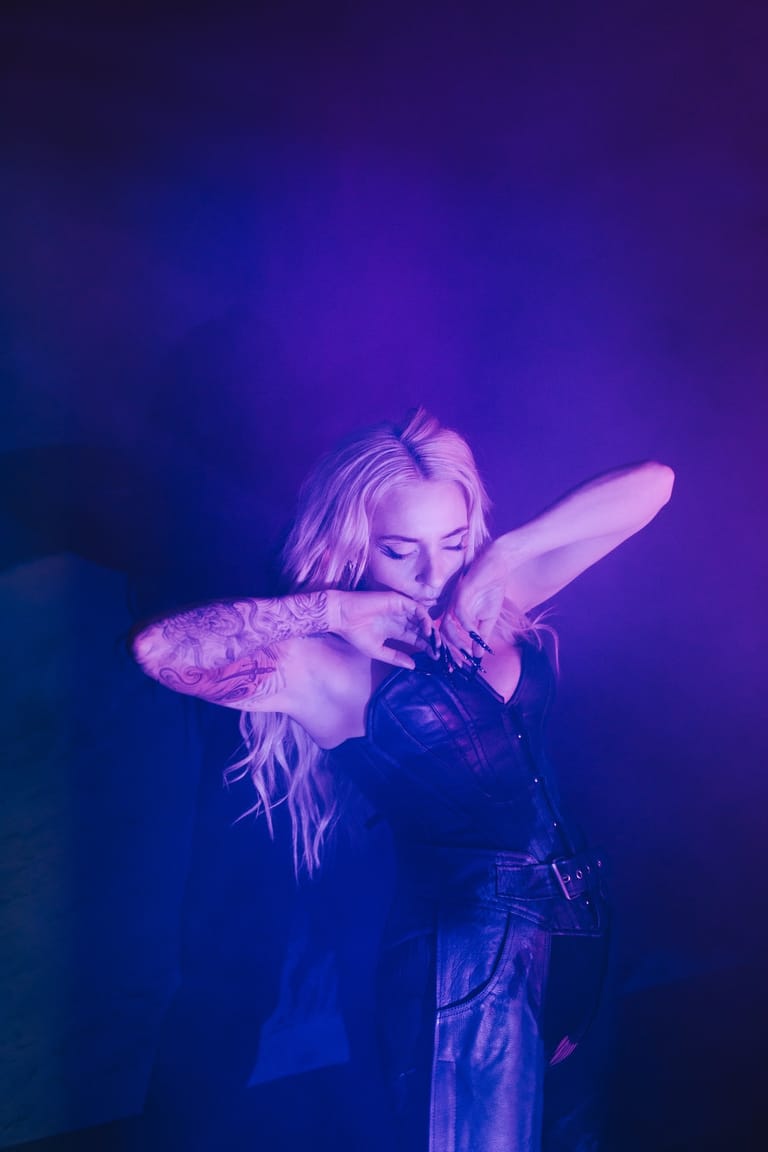 Performer with blonde hair and tattoos poses against purple stage lighting in a dark outfit.