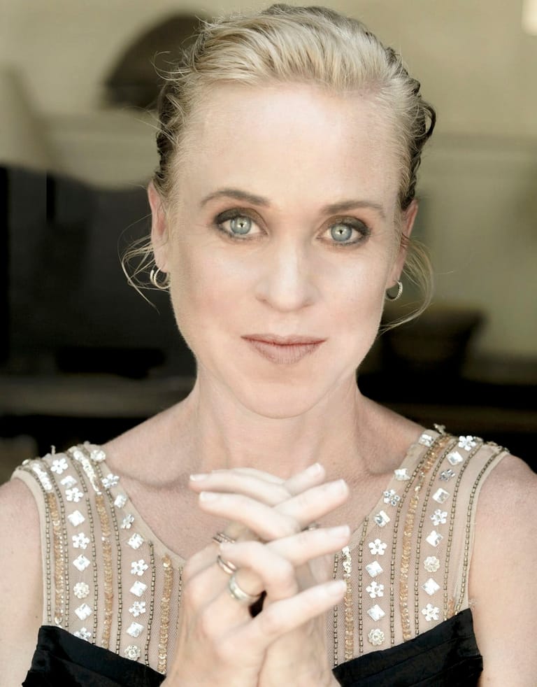 Woman with blonde hair styled back wearing an embellished beige and black sleeveless top, hands clasped at chest.