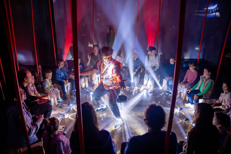 Performers surrounded by dramatic lighting and projections on stage while audience members watch from the sides.
