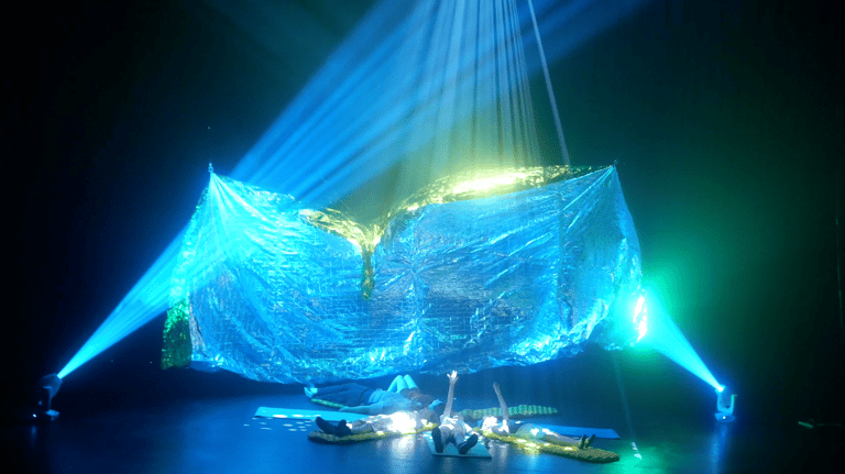Luminous blue fabric installation suspended from above, illuminated by spotlights in a darkened theatre space.
