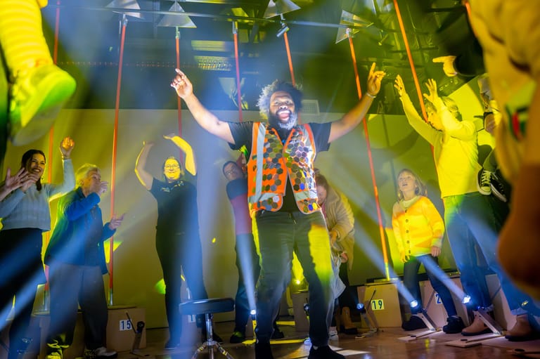 Performer in colourful jacket gestures to crowd under bright yellow and blue stage lighting.