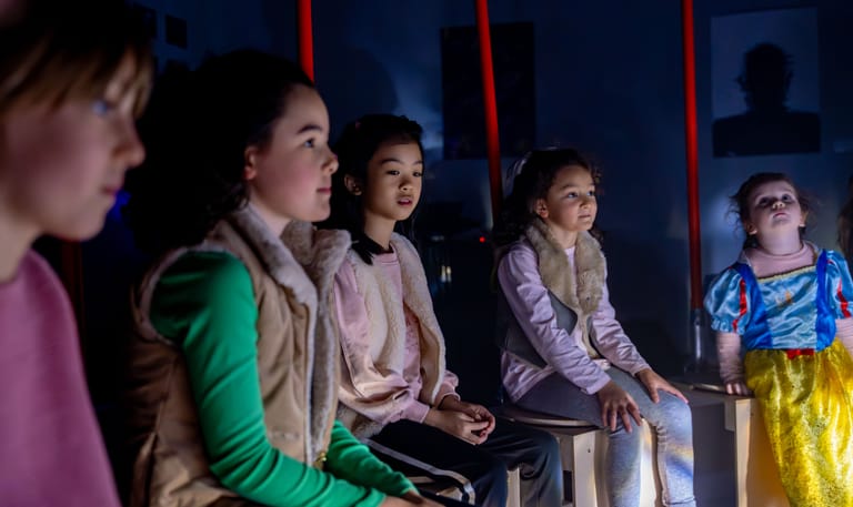Five young performers sit on a darkly lit stage beneath red suspended ropes, wearing colourful costumes.