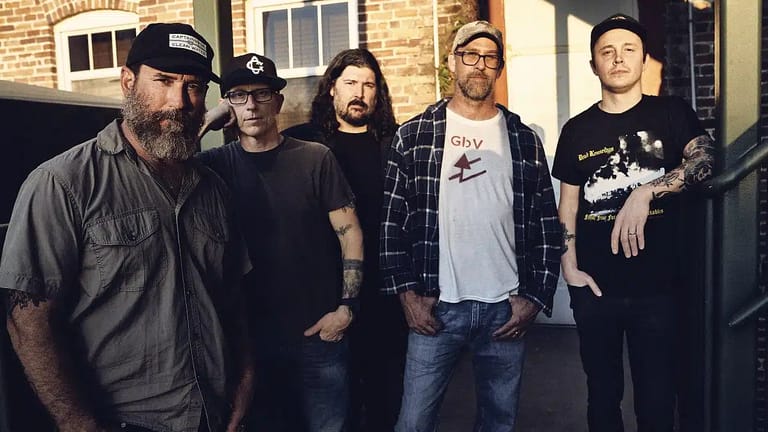Five band members posing together against a brick wall in casual streetwear and caps.