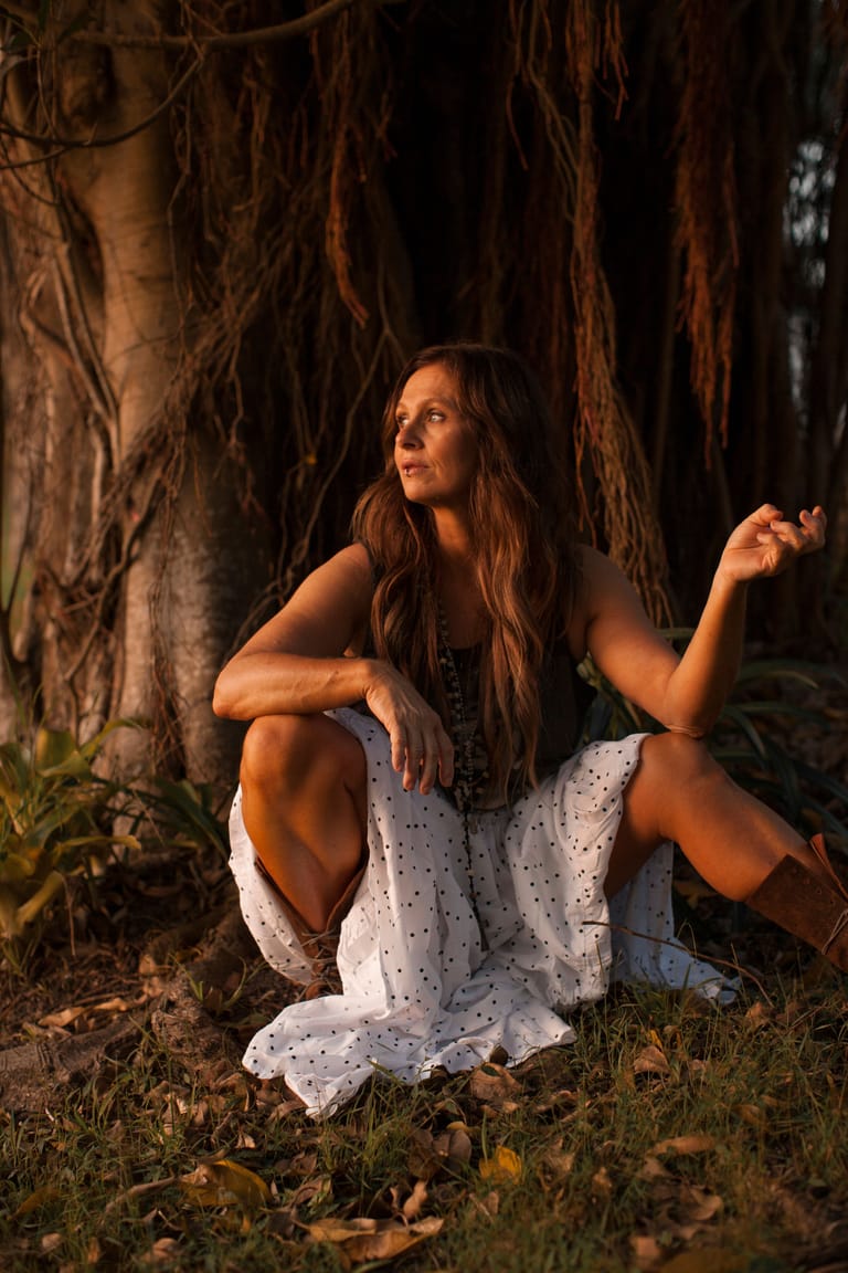 Woman in white dress crouching beneath hanging vines in golden-hour forest light.