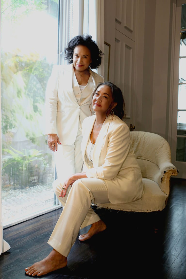 Two women in cream-coloured suits pose by a bright window in an elegant interior space.