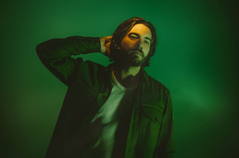 Man with hand to head against green-lit backdrop, wearing dark jacket and contemplative expression.